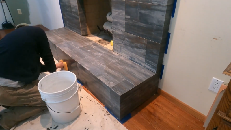 A Worker Kneels While Applying Grout Between Wood-Look Tiles on A Newly Tiled Brick Fireplace