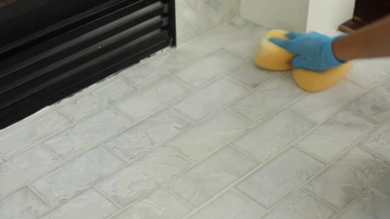 A Person Wipes Excess Grout from White Tiles on A Brick Fireplace Hearth Using a Sponge