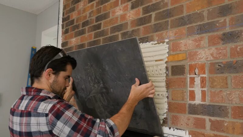 A Man Carefully Aligns a Large Dark Tile Against a Brick Fireplace, Ensuring Proper Placement Over the Fresh Adhesive