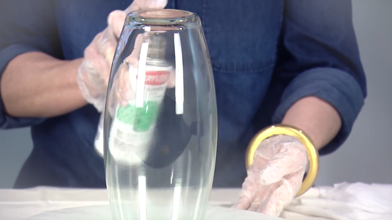 A Person Wearing Gloves Is Spray Painting a Glass Vase with Green Paint, Holding the Vase Upside Down to Ensure Even Coverage