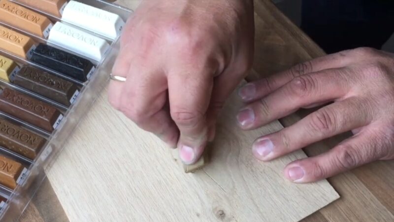 A Person Applies a Wax Stick to Fill Screw Holes in A Wooden Surface, with A Selection of Colored Wax Sticks Visible in The Background