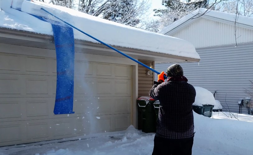 A homeowner uses a roof rake to remove accumulated snow from the edge of a residential garage roof