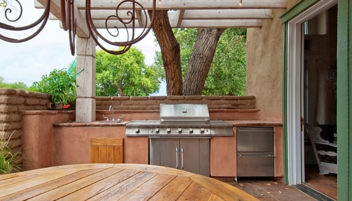 Outdoor kitchen with stainless steel grill, sink, stone countertops, wooden pergola, and dining area on a covered backyard patio.